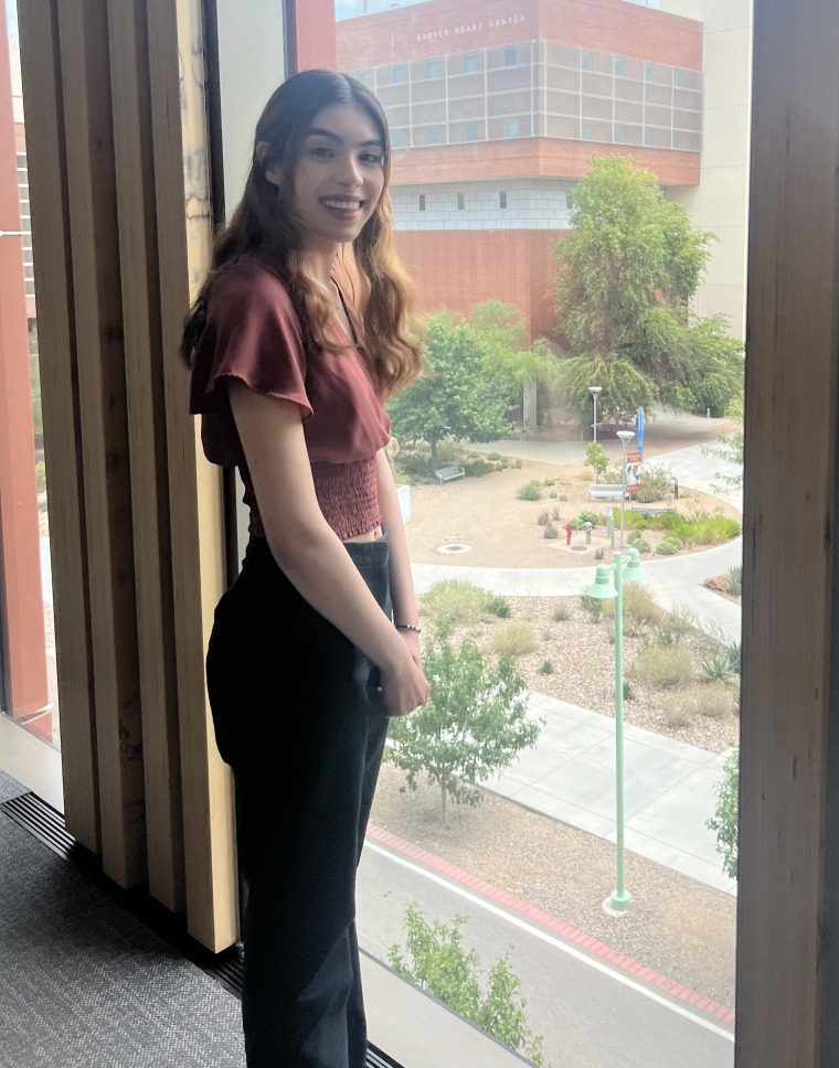 photograph of a person standing next to a window with the University of Arizona campus in the background
